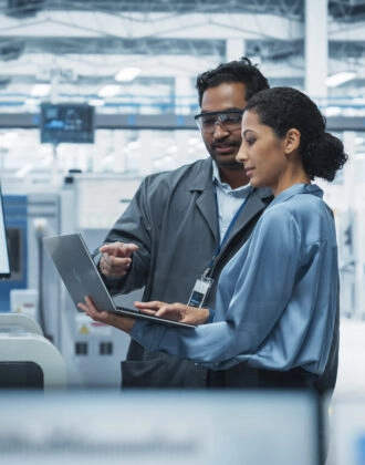 Indian Male Industrial Engineer And Hispanic Female Supervisor Using Laptop And Talking At An Electronics Factory. Man Using Soldering Jet Printer, Explaining Process Behind Production Of Motherboards